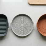 Top-down flat lay photo of three air fryer liners side by side on a clean kitchen counter: a grooved silicone basket liner, a perforated mesh silicone liner, and a solid bowl-shaped silicone liner.