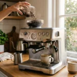 Premium coffee maker with built-in grinder (Breville Barista Express styled) on kitchen counter, whole coffee beans being poured into hopper (mid-pour), fresh cup of coffee steaming beside